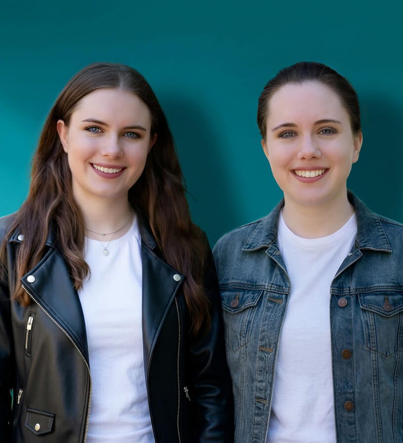 Brittany and Ashley, co-owners of Twingenuity Graphics, stand side by side in front of a teal background, both smiling at the camera.